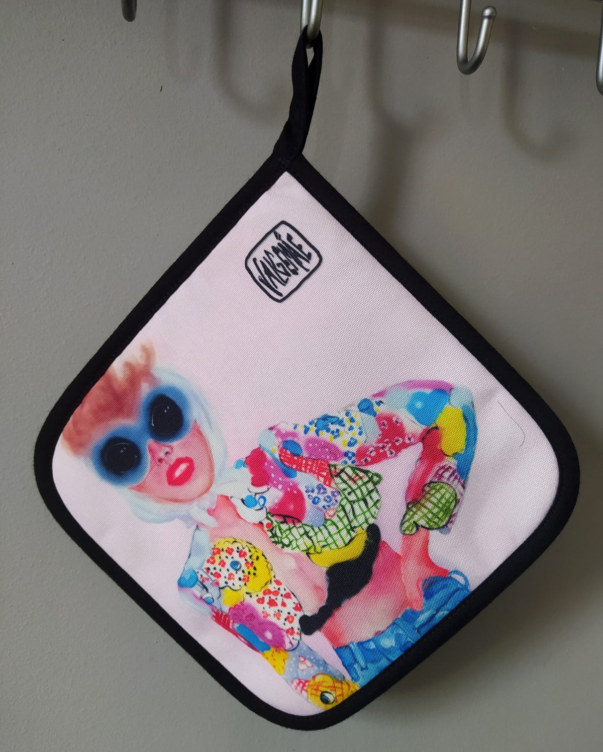 2. Pot holder with pop art design hanging on a hook, showing colorful woman illustration and Katrin Valgemäe logo