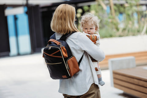 1. Woman carrying Ally Scandic Small Diaper Backpack in Black Coffee while holding a child outdoors
