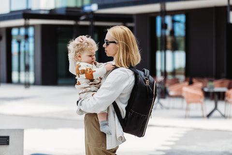1. Woman carrying Ally Scandic Espresso Diaper Backpack while holding a child, showcasing hands-free convenience