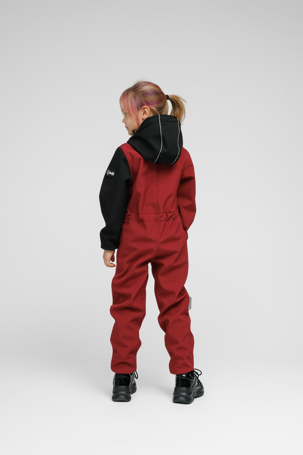 5. Back view of child in Nuckö sunset red softshell onesie with black hood, highlighting reflective stripe and elastic waist