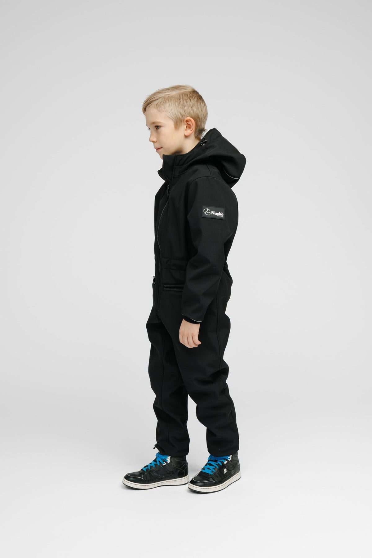 5. Side view of child in black softshell onesie with hood and visible logo on sleeve in studio