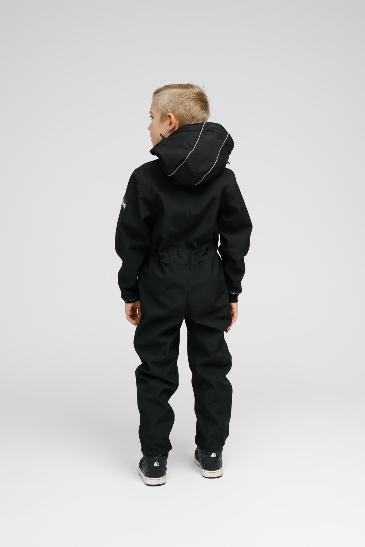 3. Back view of child in black softshell onesie with hood and blue sneakers in studio