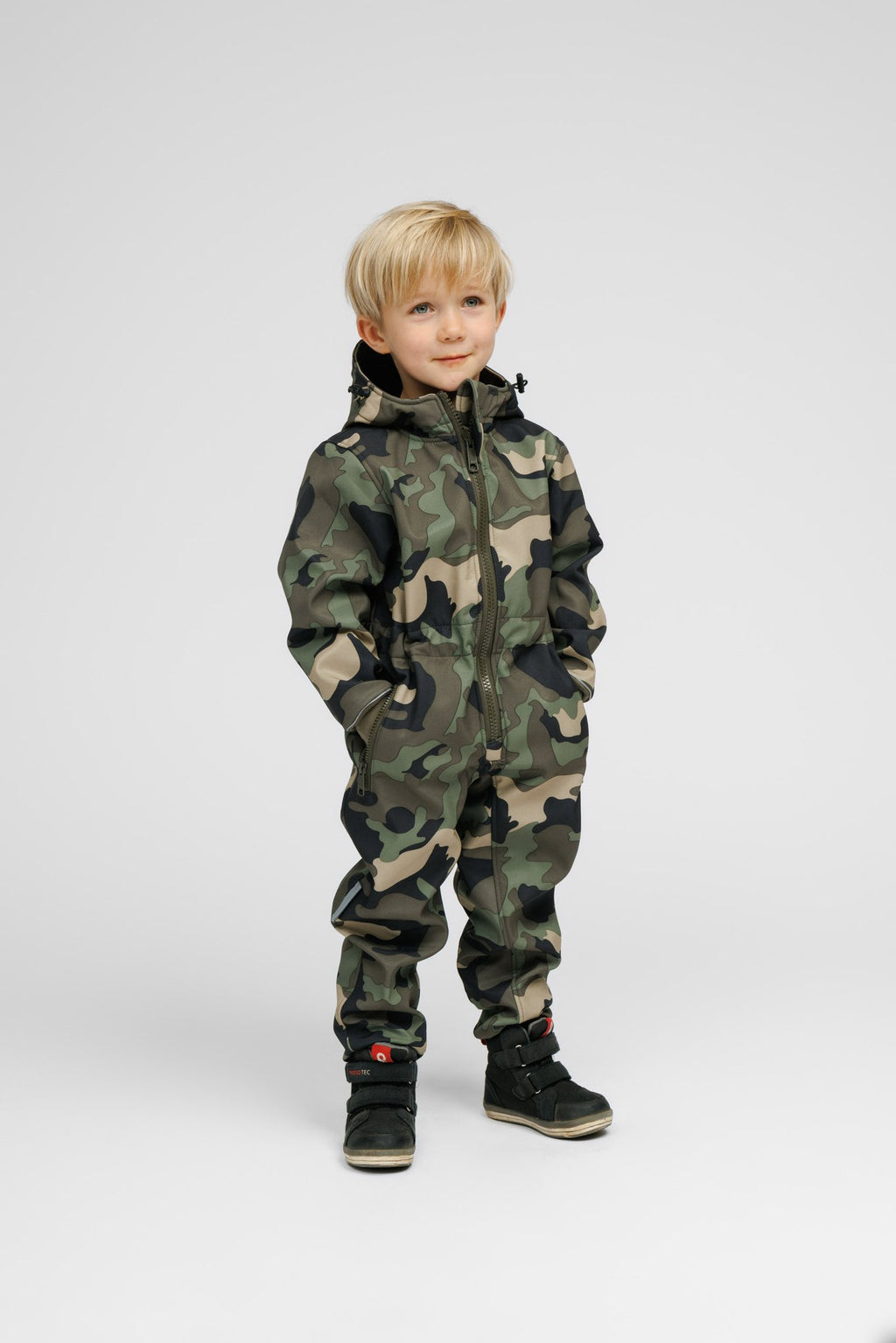 1. Child wearing Nuckö softshell onesie in camouflage pattern, standing confidently with hands in pockets, showcasing adjustable hood and ribbed cuffs, studio background