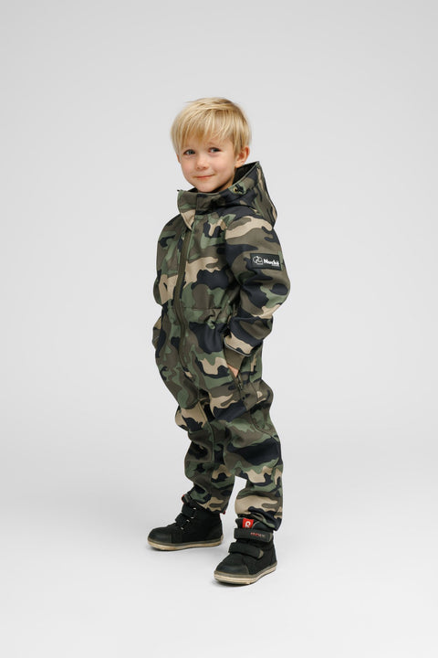 4. Child in Nuckö camouflage softshell onesie, smiling with hands in pockets, displaying brand logo on sleeve, studio background