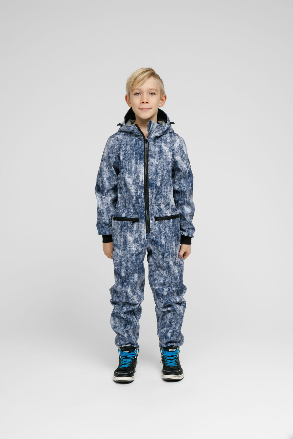 1. Child wearing blue patterned softshell onesie with hood and black sneakers standing in studio