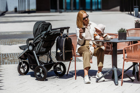 2. Woman sitting at a cafe with Ally Scandic Espresso Diaper Backpack attached to stroller, highlighting practical use