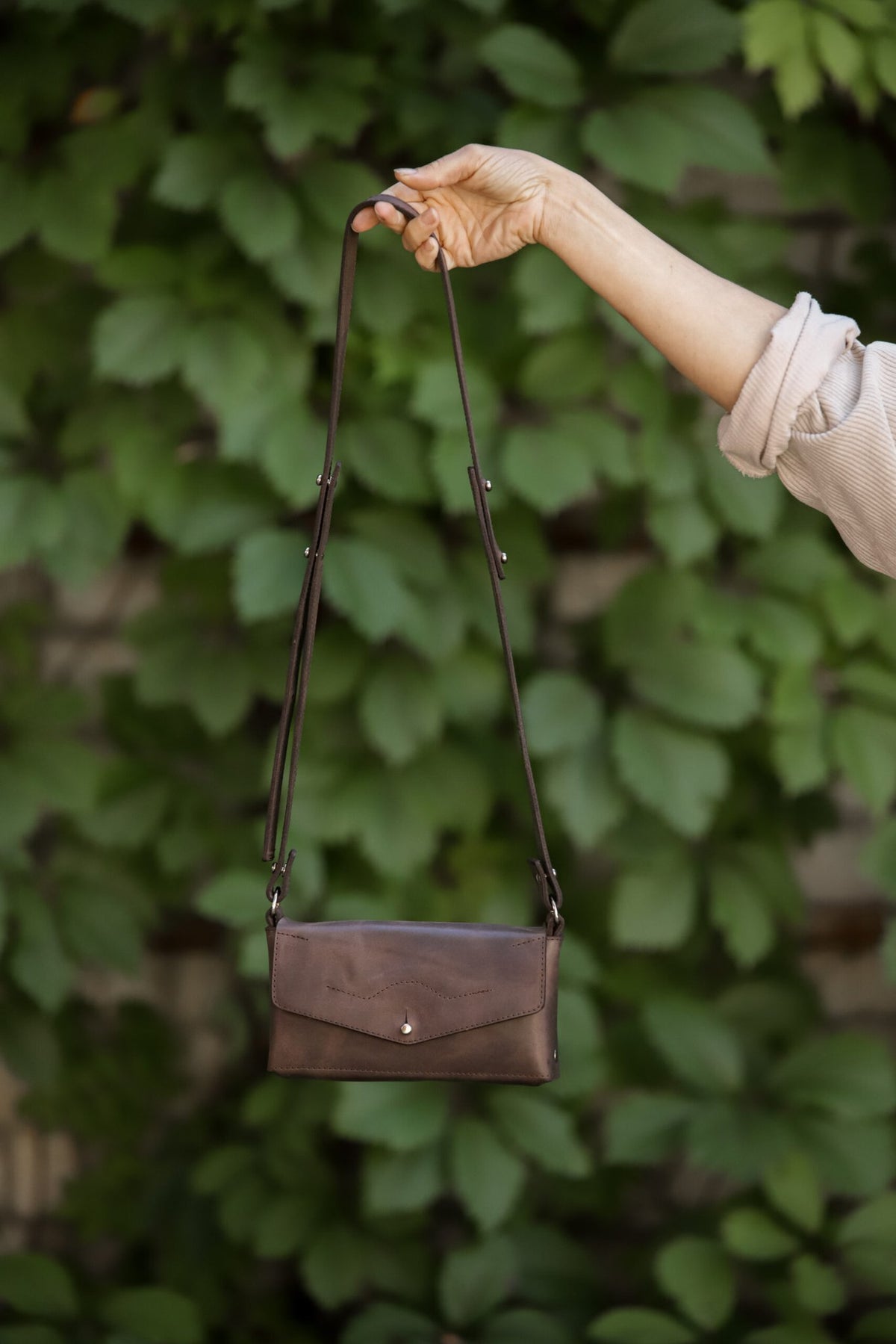 8. KOKUKA crossbody leather bag in brown held by hand against a leafy background, showcasing design