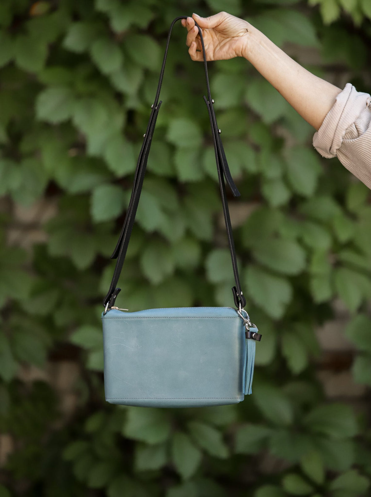 3. Lifestyle shot of eleriinseim TOURIST crossbody leather bag in Capri blue, held by a woman against a leafy background