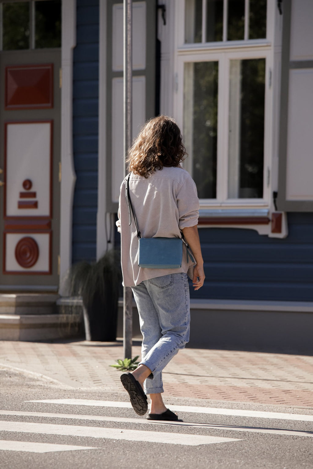 1. Woman walking with eleriinseim TOURIST crossbody leather bag in Capri blue, showcasing adjustable strap and compact design