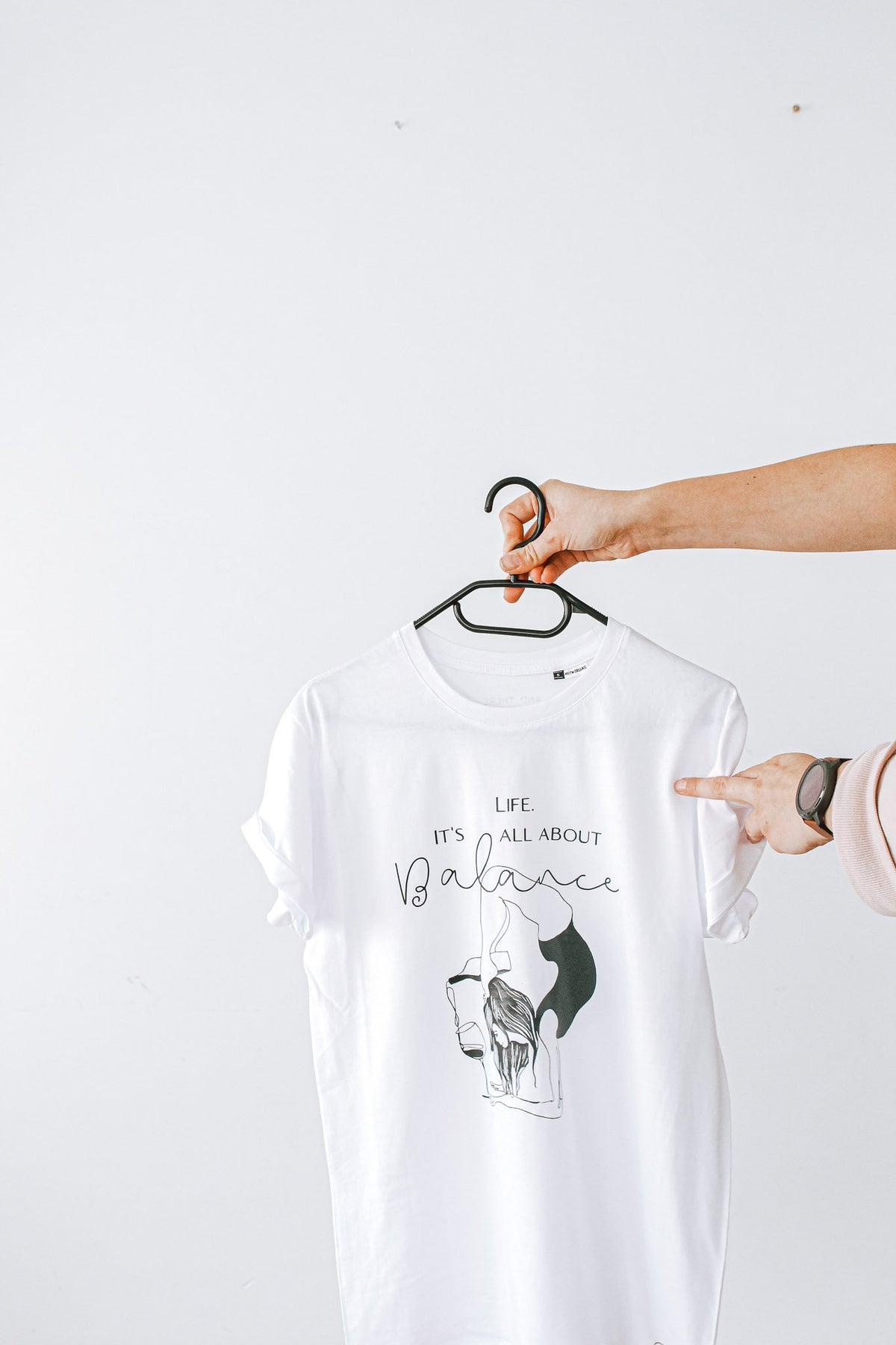 4. Close-up of white 'It's All About Balance' T-shirt by Black Giraffe Brand, held on hanger with rolled-up sleeves