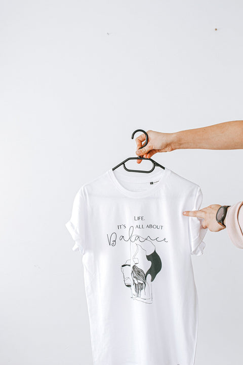 4. Close-up of white 'It's All About Balance' T-shirt by Black Giraffe Brand, held on hanger with rolled-up sleeves