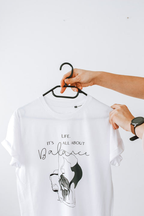 3. Close-up of white 'It's All About Balance' T-shirt by Black Giraffe Brand on hanger, highlighting design and rolled-up sleeves