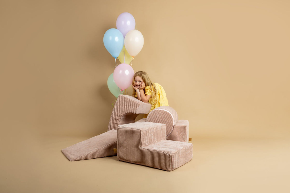 1. Girl in yellow dress leaning on powder pink shape set with balloons in a beige room