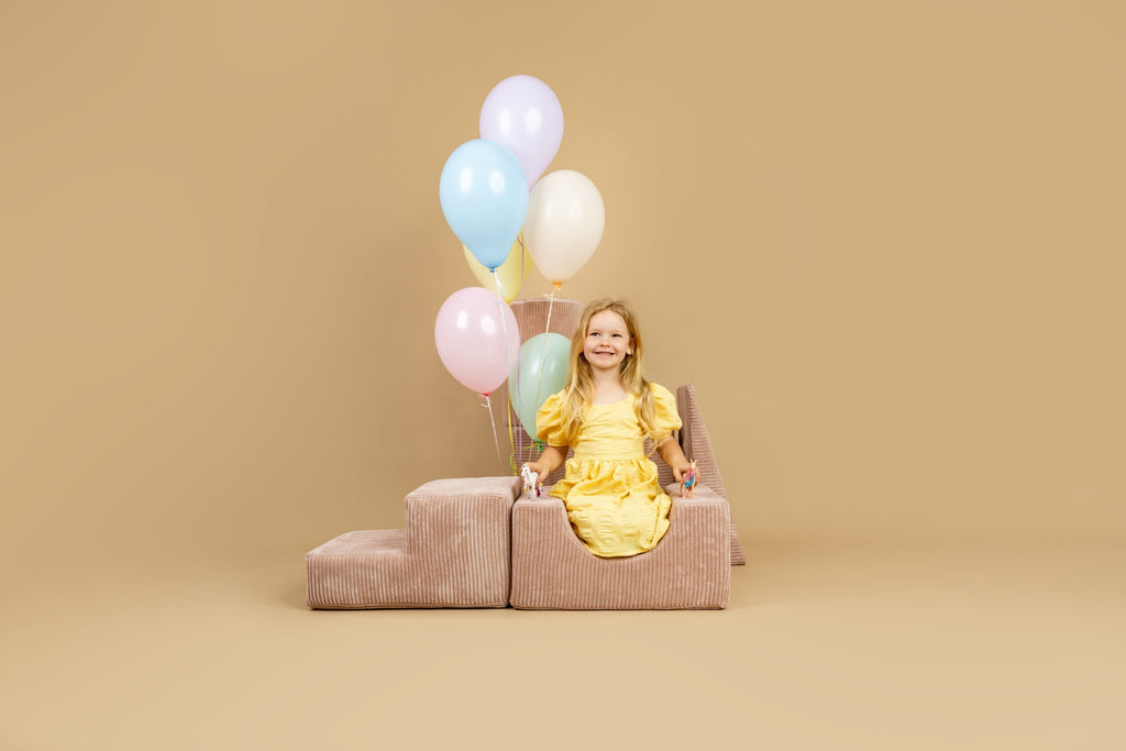1. Smiling girl in yellow dress sitting on powder pink shape set with pastel balloons, in a cheerful indoor environment