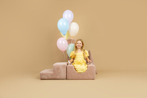 1. Smiling girl in yellow dress sitting on powder pink shape set with pastel balloons, in a cheerful indoor environment