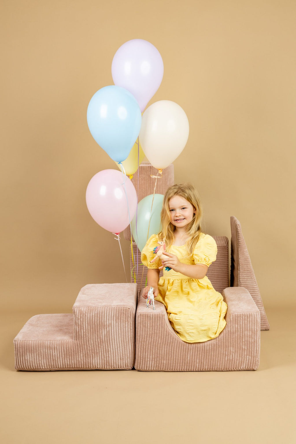 1. Young girl in yellow dress playing with powder pink shape set, holding colorful balloons in a playful indoor setting