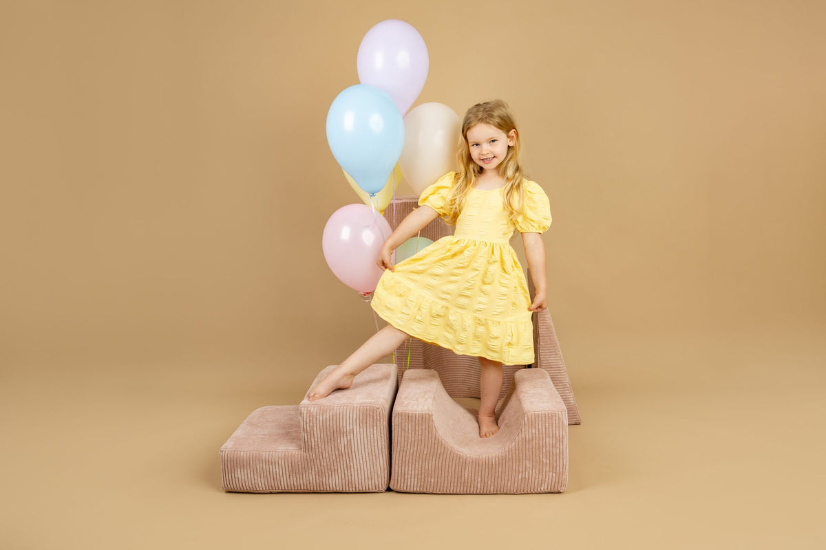 1. Girl in yellow dress standing on powder pink shape set, holding balloons, in a joyful indoor setting