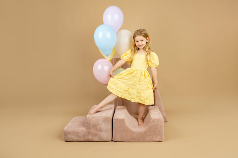1. Girl in yellow dress standing on powder pink shape set, holding balloons, in a joyful indoor setting