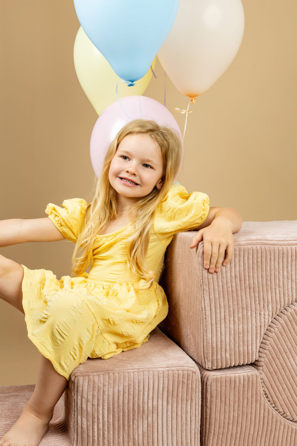 1. Girl in yellow dress sitting on powder pink shape set with balloons in a beige room