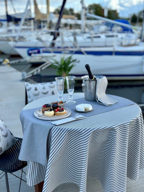 4. Outdoor dining scene with Hortensias Home round tablecloth in beige with blue lines, set with desserts and champagne by the marina