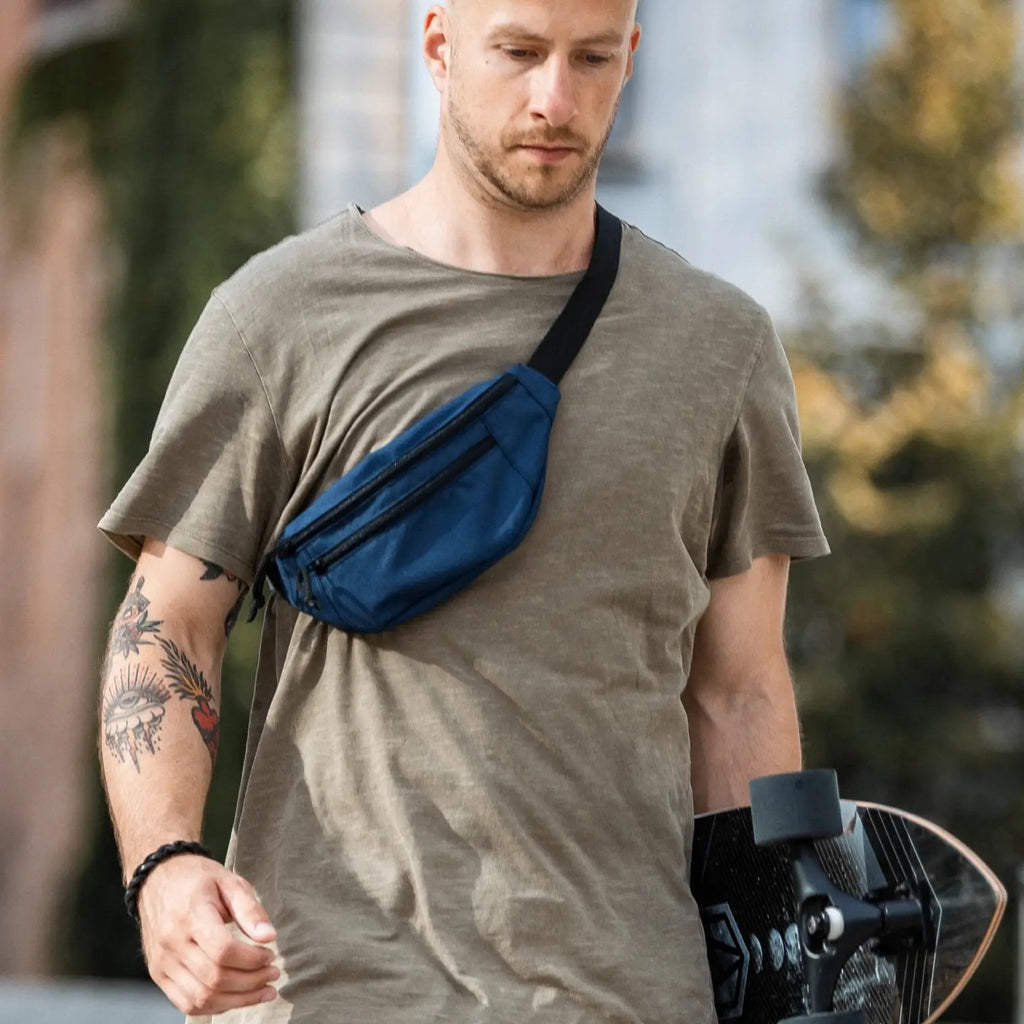 1. Men's blue Drinbags waist bag worn crossbody, shown in lifestyle setting with skateboard