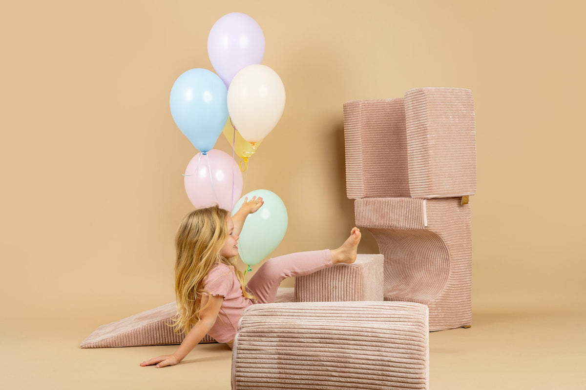 1. Girl in pink outfit reaching for balloons while sitting on powder pink shape set in a beige room