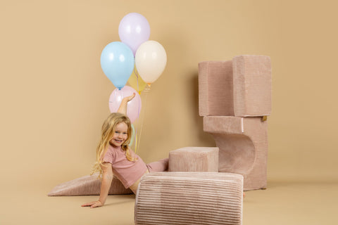 1. Smiling girl in pink outfit with balloons sitting on powder pink shape set in a beige room