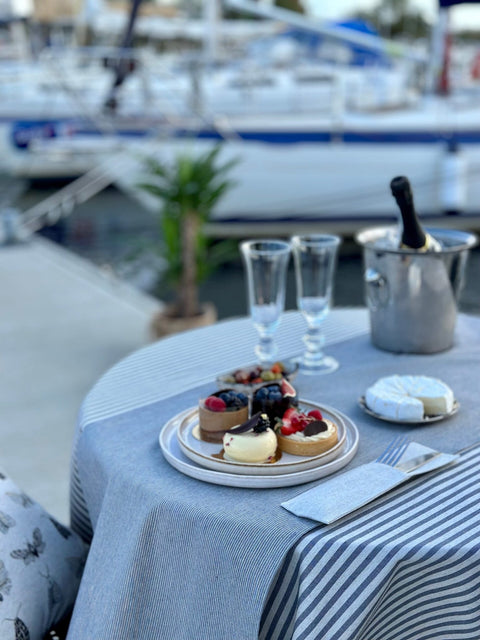 2. Close-up of Hortensias Home Nord 2 table runner with blue stripes, featuring desserts and champagne glasses, set on a table by the marina