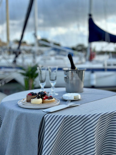 1. Lifestyle image of Hortensias Home Nord 2 table runner with blue stripes, styled on a dining table with desserts and champagne, set outdoors by a marina