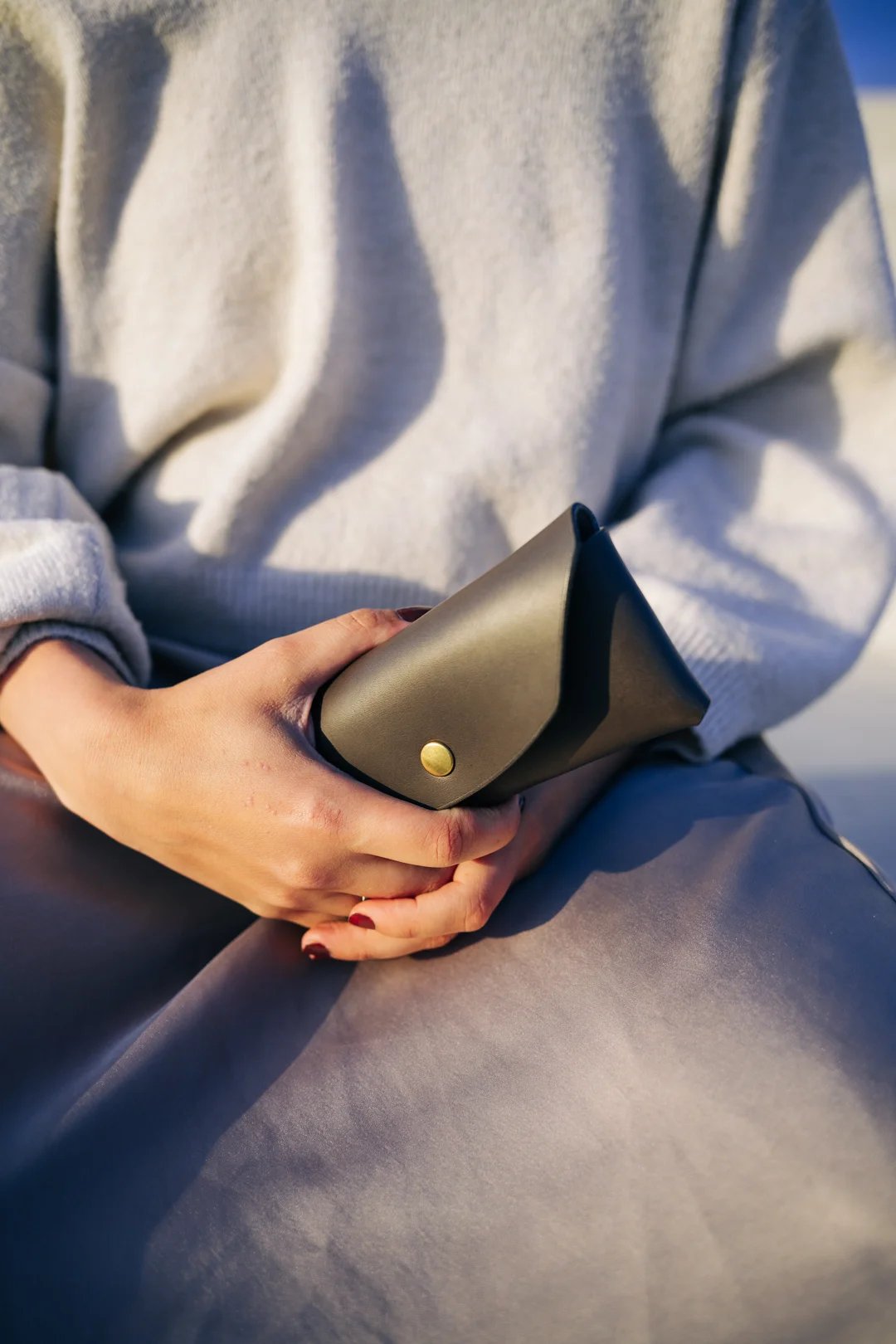 1. Woman holding Mirgoods black leather sunglasses case with gold button closure, lifestyle setting