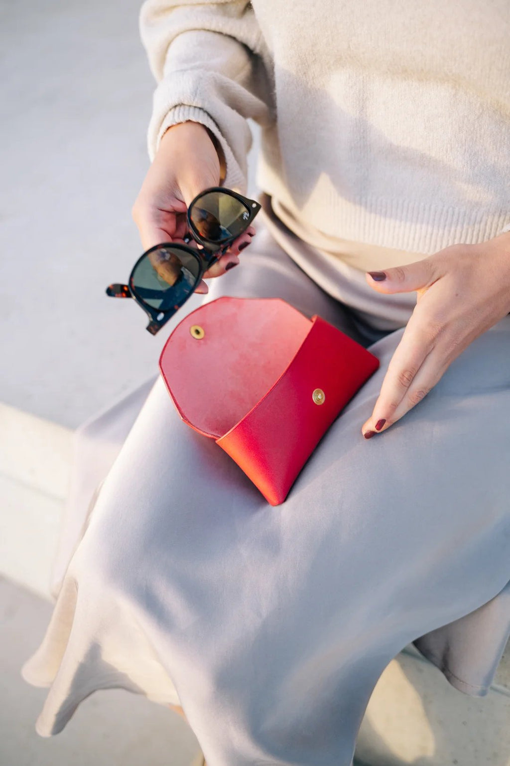 2. Woman placing sunglasses into Mirgoods red leather case on her lap