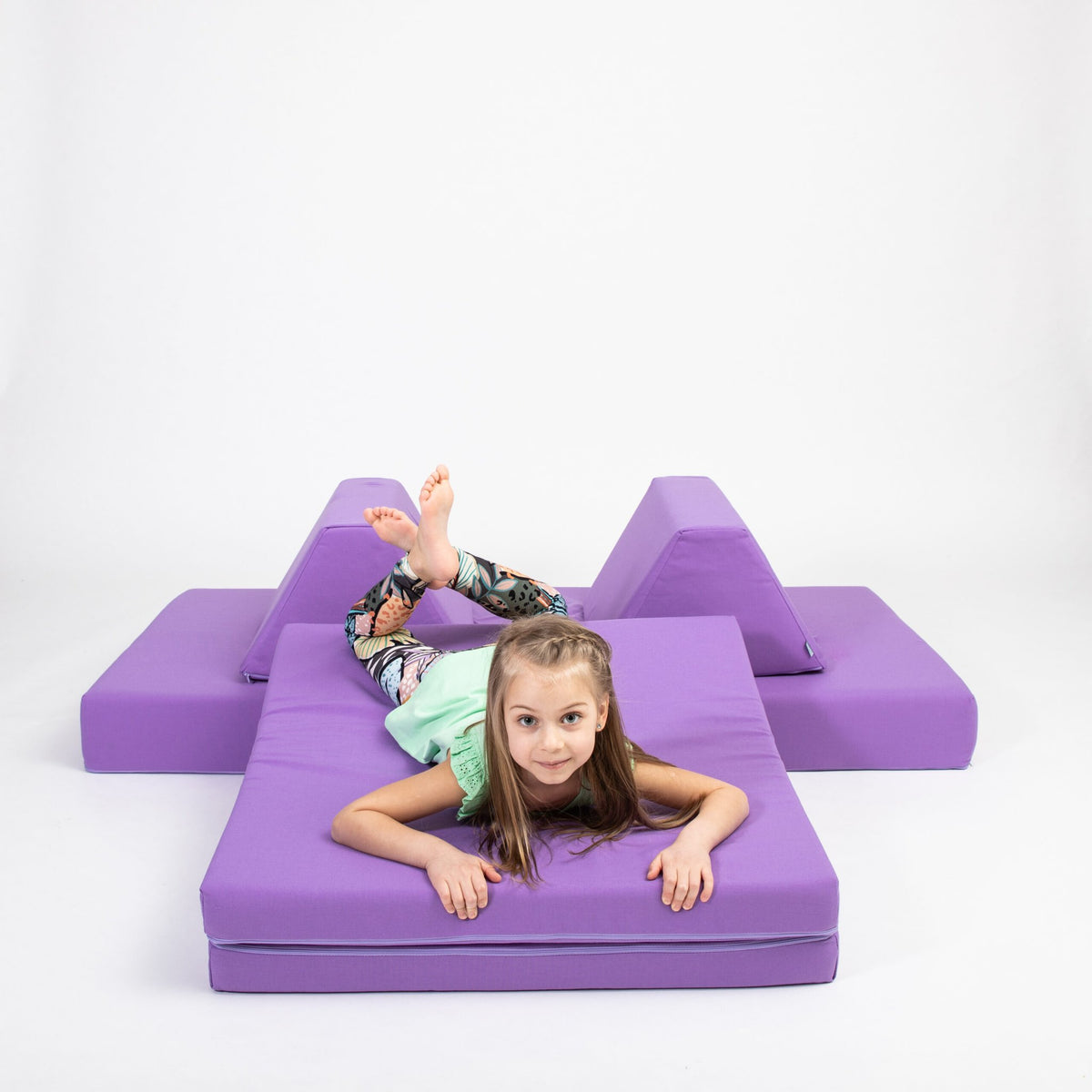 1. Young girl lying on purple activity play mattress set with trapezoidal pillows in a playful pose on a white background