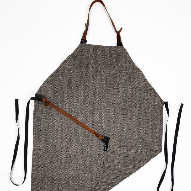 3. Studio shot of KOOSdisain black and gray herringbone linen apron with leather belt and pocket, laid flat showing full design