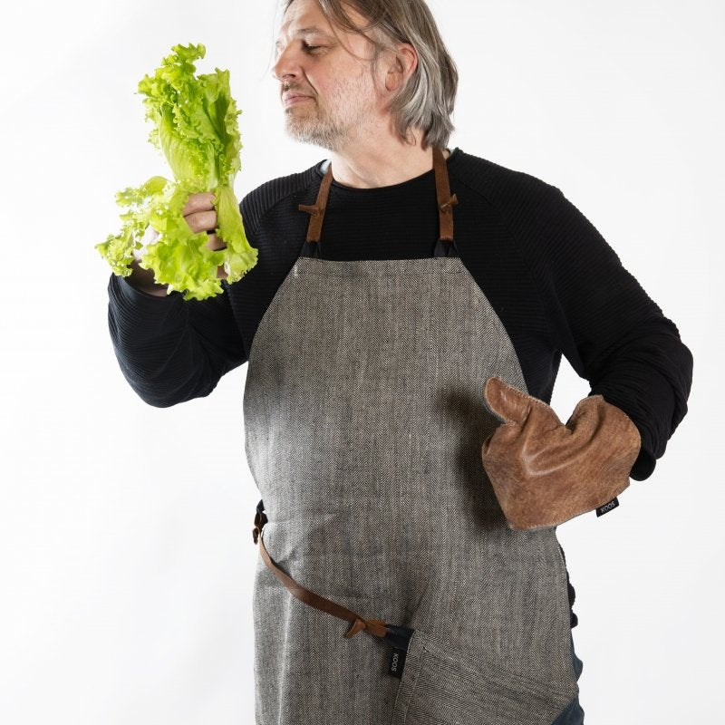 1. Man wearing KOOSdisain black and gray herringbone linen apron with leather details, holding lettuce and wearing a brown oven mitt in a studio setting
