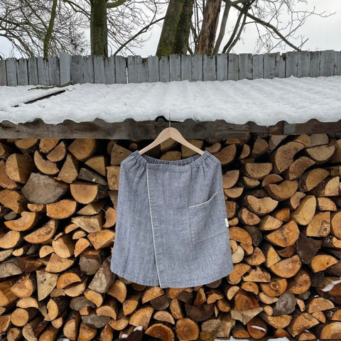 1. Grayish blue linen sauna skirt with fishbone pattern hanging on wooden hanger against stacked logs outdoors