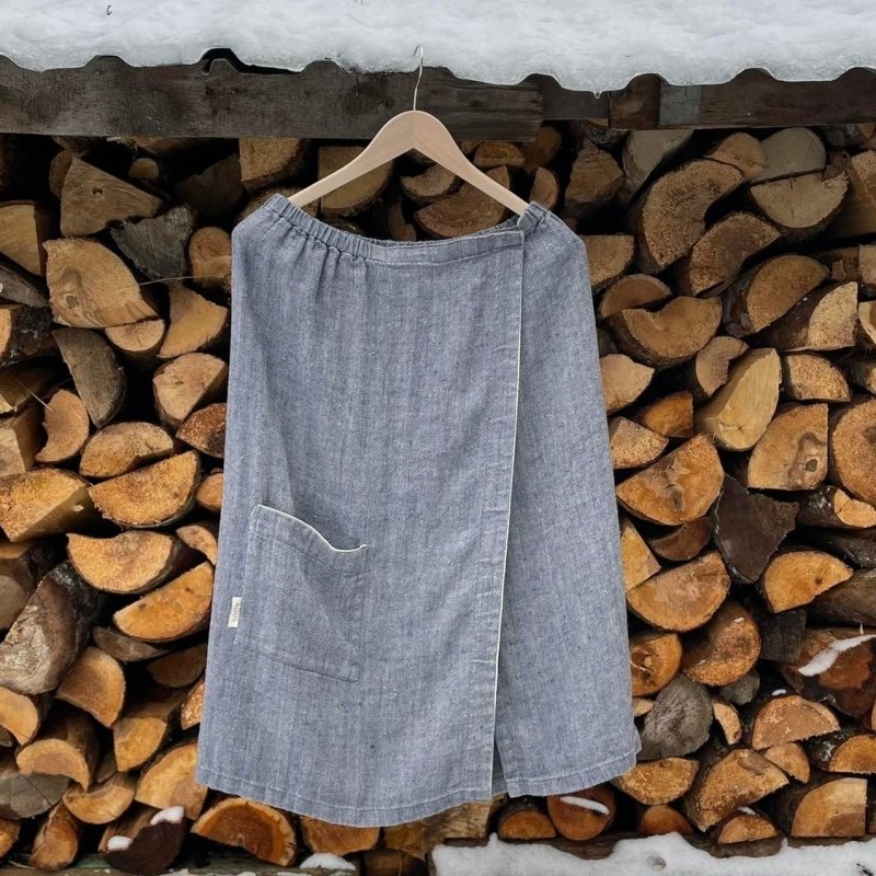 1. Women's grayish blue linen sauna skirt with fishbone pattern hanging on a wooden hanger against a stack of firewood