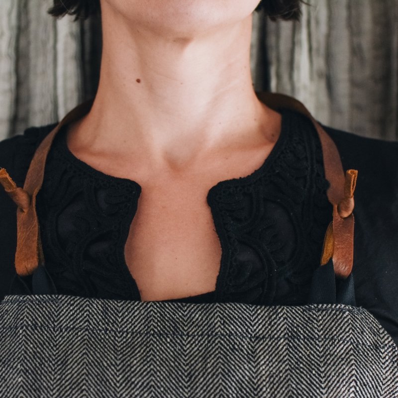 5. Close-up of KOOSdisain apron showing leather strap detail on black and gray herringbone fabric worn by a woman