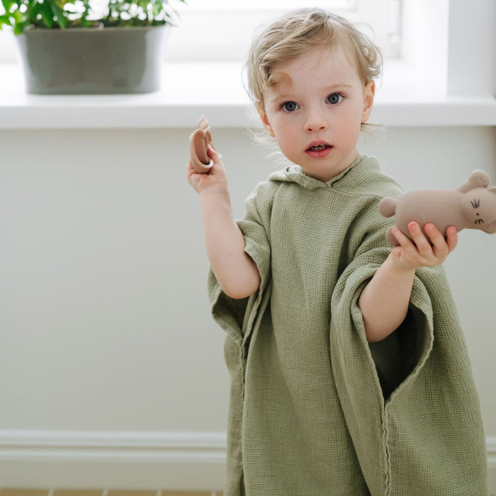 1. Child wearing sage green linen poncho with hood, holding toys, standing indoors near a window