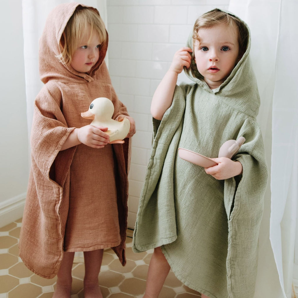 1. Two children wearing linen ponchos, one in sage green and the other in peach, standing in a bathroom holding toys