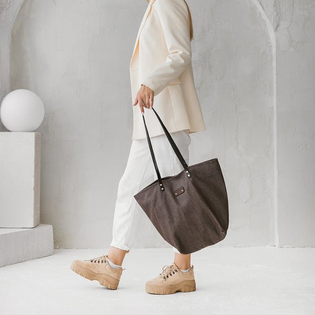 4. Side view of woman holding Muni brown denim tote bag with black leather handles in a lifestyle setting