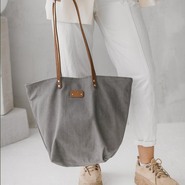 1. Women's grey denim tote bag by Muni with tan leather handles, styled with white pants and beige shoes in lifestyle setting