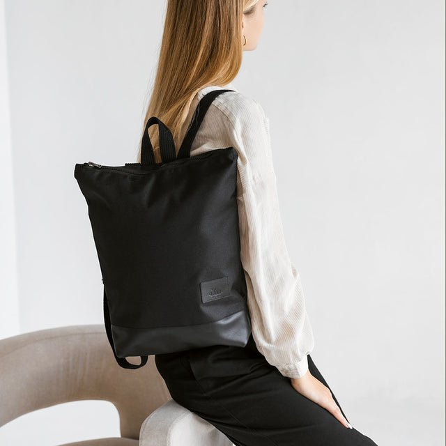 1. Woman sitting with Muni waterproof backpack in black leather and polyester, showcasing adjustable straps and sleek design