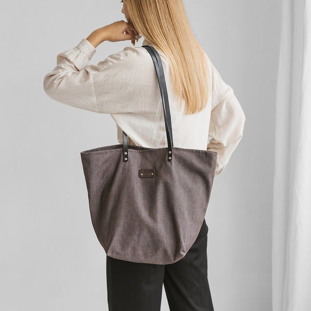 1. Woman carrying Muni brown denim tote bag with black leather handles, shown from the back in a lifestyle setting