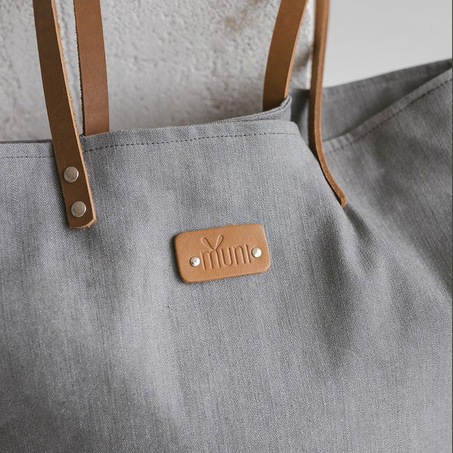 9. Close-up of Muni grey denim tote bag, highlighting tan leather handles and brand logo with metal rivets