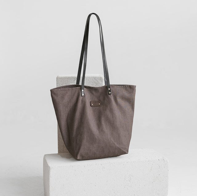 2. Muni brown denim tote bag with black leather handles displayed on a white block in a studio setting