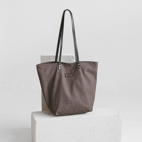 2. Muni brown denim tote bag with black leather handles displayed on a white block in a studio setting