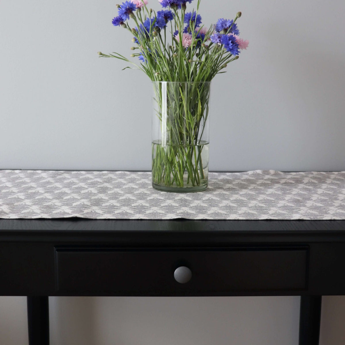 1. Table runner with Hanikatsi fly pattern on dark table, featuring a vase of blue and pink flowers, showcasing linen-cotton blend fabric