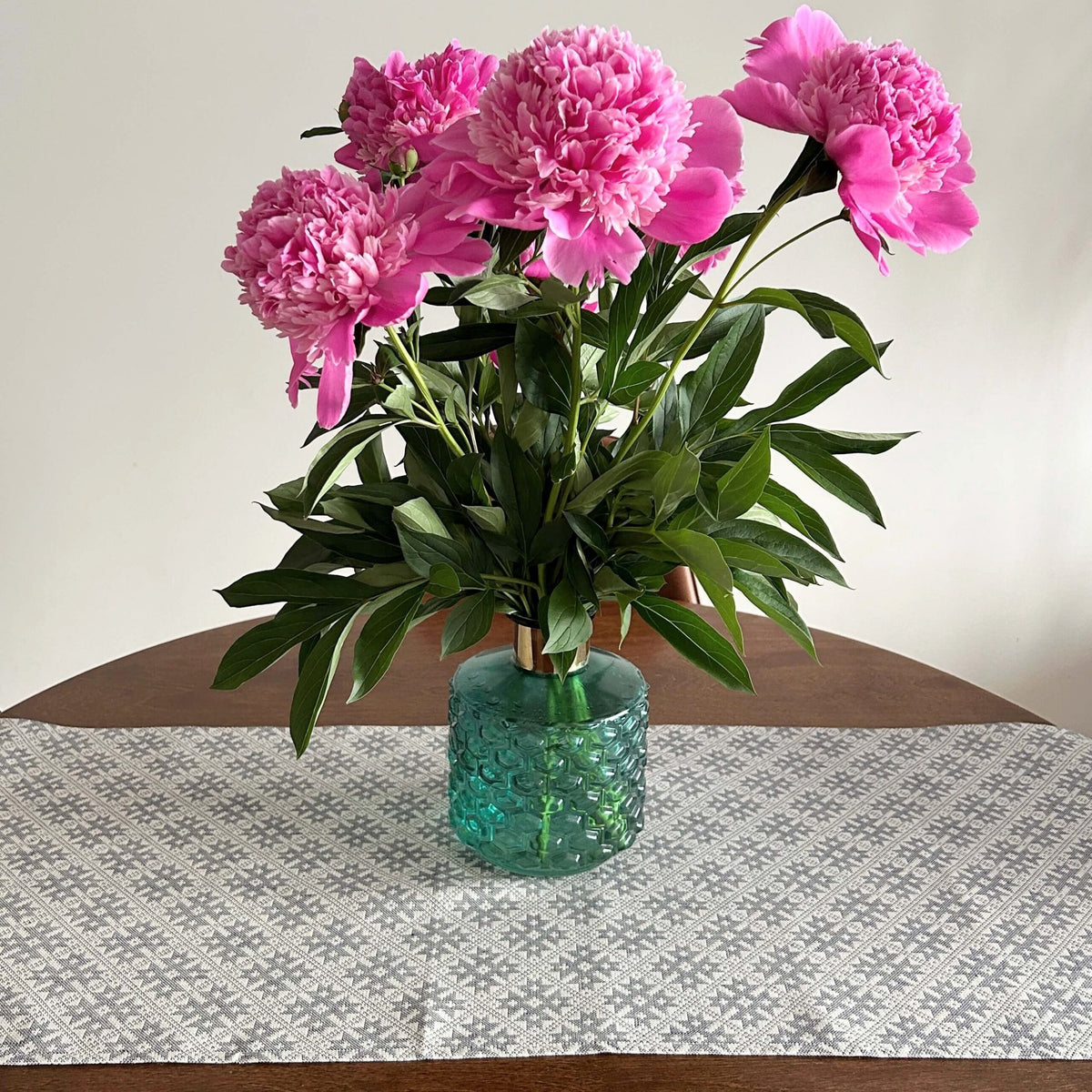 1. Grey table runner with Pärnumaa folk art pattern on wooden table, featuring a green vase with pink peonies
