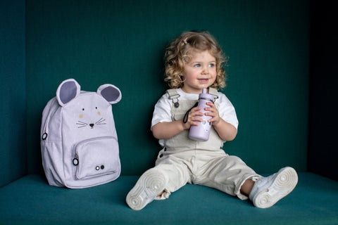 1. Child holding Trixie Baby Mrs Mouse stainless steel bottle sitting next to matching mouse backpack on a teal background