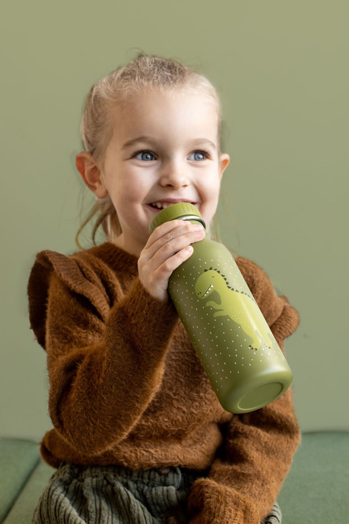 1. Young girl holding green stainless steel bottle with dinosaur print, smiling indoors